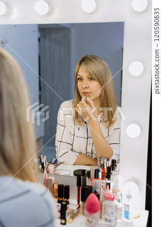 Reflection of a woman applying lipstick in a makeup mirror with beauty products on the table 120590635