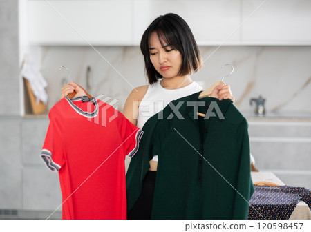 Sad young Asian girl trying to choose a dress in the kitchen 120598457