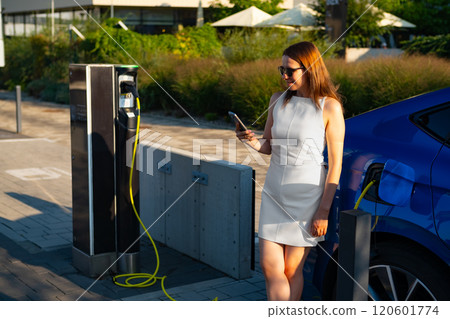 Woman stands beside her blue electric car, using her mobile phone at a charging station outdoors 120601774