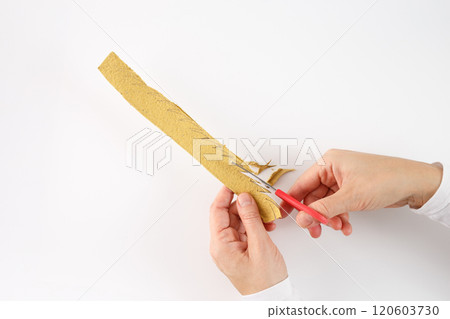 a pair of hands holding a strip of yellowish material, egg box carton, and using a pair of red-handled scissors to cut it. a zigzag pattern drawn on it, which is being followed by the scissors 120603730