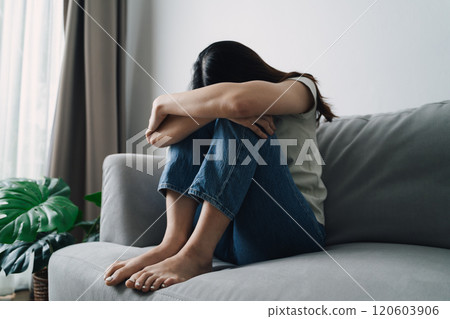 A depressed woman is sitting on a couch with her head down. She is sad or upset. 120603906