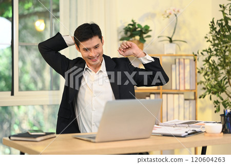 Smiling adult businessman stretching his arms while sitting at desk relaxing during a busy workday Smiling adult businessman stretching his arms while sitting at desk relaxing during a busy workday 120604263