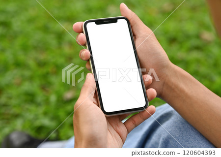 Close up of hands holding a smartphone with blank screen on background of green grass 120604393