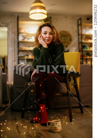 A cheerful woman in green knit sweater and red socks sits in cozy home interior, holding festive mug and surrounded by glowing garland lights, creating a warm and joyful Christmas, New Year atmosphere 120606232