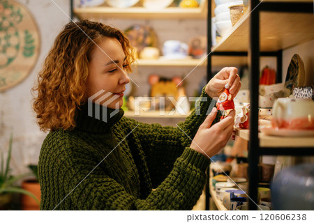 A cheerful woman in a green knit sweater selects a handmade ceramic Christmas decoration in a festive artisan shop. The image highlights the charm of local craftsmanship and holiday spirit 120606238