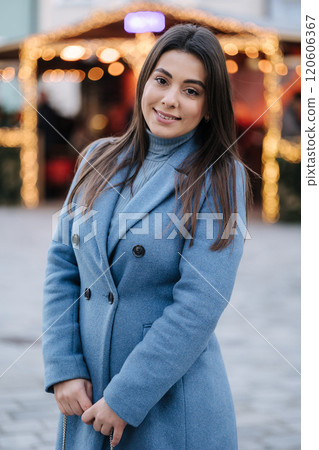 Portrait of happy beautiful female in blue winter coat standing in front of Christmas light in old town. Winter mood 120606367