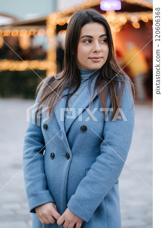 Portrait of happy beautiful female in blue winter coat standing in front of Christmas light in old town. Winter mood 120606368