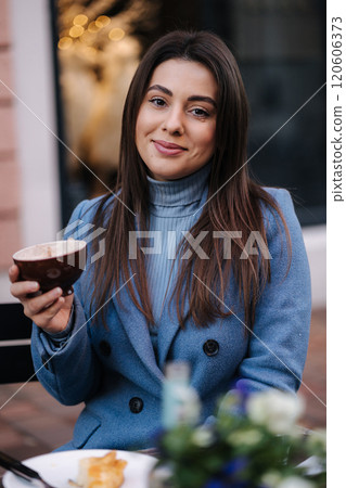 Young beautiful woman sitting outdoor in cafe and drinking coffee. Happy female in blue winter coat. Christmas mood 120606373