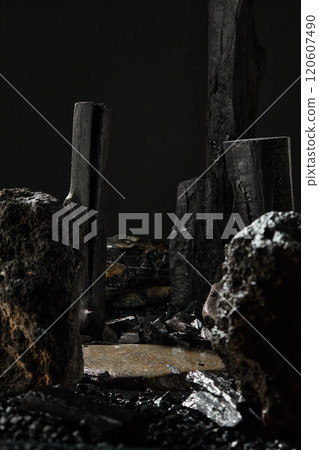 In the dark background, a close-up view of a black stone plate with an empty space to display the product, all around are some charcoal sticks and stone blocks on the pebble surface. 120607490