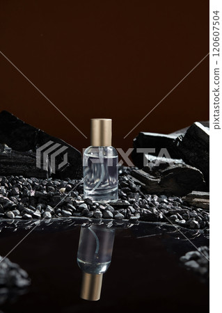 In the brown backdrop, a frontal view of a glass bottle with a silver cap, containing a liquid, is standing on a dark pebbles surface and has a reflection below, behind are some charcoal blocks. In the brown backdrop, a frontal view of a glass bottle with a silver cap, containing a liquid, is standing on a dark pebbles surface and has a reflection below, behind are some charcoal blocks. 120607504
