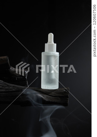 An opaque white glass serum bottle placed on the wooden coal in the center of the frame. Black background with the same smoke flying. A minimalist photo template, perfect for cosmetic advertising. 120607506