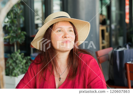 Millennial woman in a straw hat and red blouse sits at an outdoor cafe, gazing thoughtfully into the distance. Concept of relaxation, summer mood and style and fashion Millennial woman in a straw hat and red blouse sits at an outdoor cafe, gazing thoughtfully into the distance. Concept of relaxation, summer mood and style and fashion 120607863