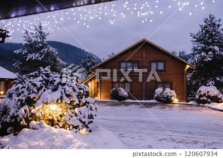 Charming wooden house entrance lit by festive lights, snow-covered stairs, and peaceful winter surroundings. 120607934