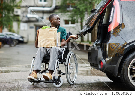 Full length scene with of young African American man using wheelchair opening car trunk in parking lot and holding groceries while doing shopping in city 120608166