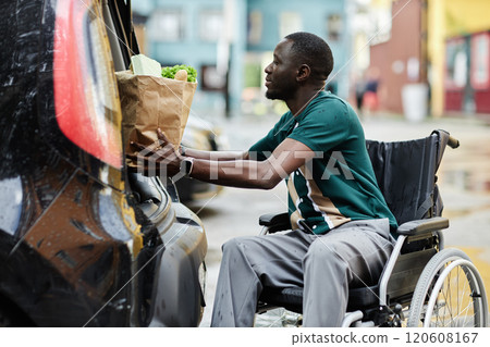 Side view portrait of young African American man with disability loading in groceries in car trunk while doing shopping in city 120608167