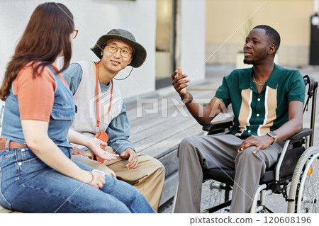 Diverse group of three adult friends with African American man using wheelchair chatting happily outdoors in city Diverse group of three adult friends with African American man using wheelchair chatting happily outdoors in city 120608196