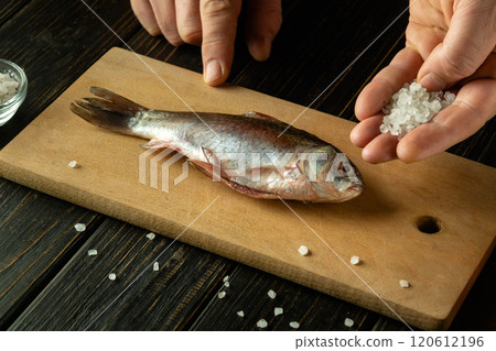 Hands sprinkle salt over a whole fish laid on a rustic wooden board. The kitchen exudes warmth, with a focus on the process of seasoning the fresh catch, ready for cooking. 120612196