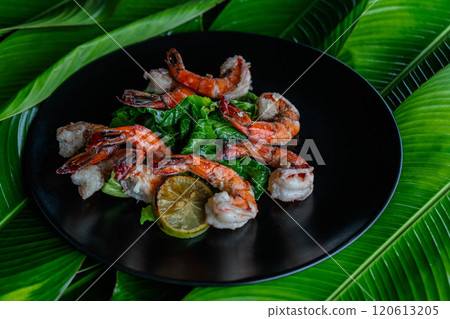 Exquisite tiger shrimp arranged elegantly on a dark plate, complemented by fresh greens and a slice of lime, showcase a vibrant Asian culinary tradition amidst lush banana leaves. 120613205