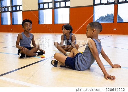 In school gym, three multiracial boys sitting on floor, wearing sports uniforms, talking 120614212