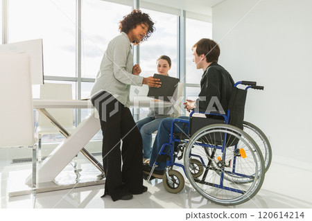 Teamwork brainstorm. Diverse multiracial colleagues man in chair for people with disability working together. Group of people sitting in creative office discussing business. Young professional team Teamwork brainstorm. Diverse multiracial colleagues man in chair for people with disability working together. Group of people sitting in creative office discussing business. Young professional team 120614214