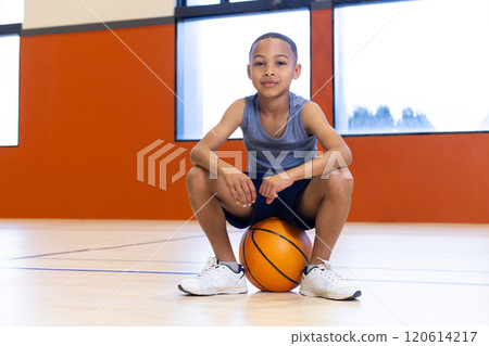 In school gym, boy sitting on basketball, looking confident and relaxed 120614217