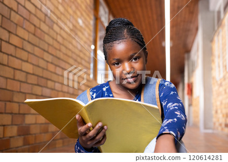 Holding notebook, african american girl smiling and reading in school hallway, wearing backpack Holding notebook, african american girl smiling and reading in school hallway, wearing backpack 120614281