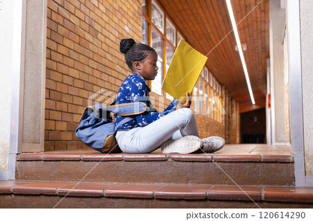 In school, african american girl sitting on steps reading yellow folder with backpack nearby 120614290