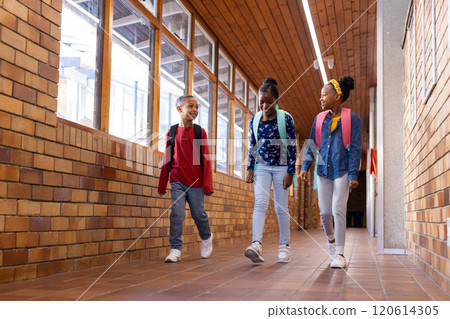 Walking in school hallway, two multiracial girls and boy with backpacks talking and smiling 120614305