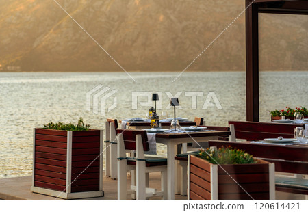 An elegant terrace cafe by the bay, featuring stylish tables and scenic views of mountains and water during a serene sunset. An elegant terrace cafe by the bay, featuring stylish tables and scenic views of mountains and water during a serene sunset. 120614421