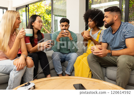 Diverse friends sitting on couch, drinking coffee, and chatting in cozy living room 120614592
