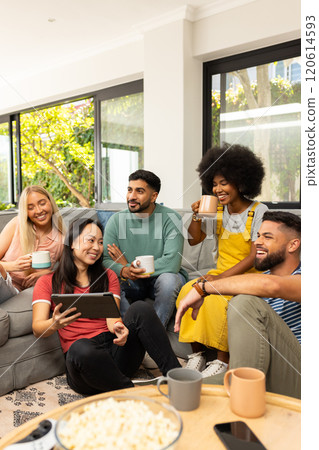 Diverse friends sitting on couch, drinking coffee, and watching tablet together, smiling 120614593
