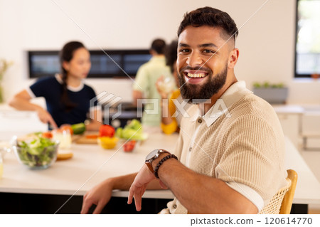 Smiling man sitting in kitchen while diverse friends preparing food in background 120614770