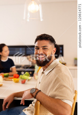 Smiling man sitting at kitchen counter with diverse friends preparing food in background, copy space Smiling man sitting at kitchen counter with diverse friends preparing food in background, copy space 120614771