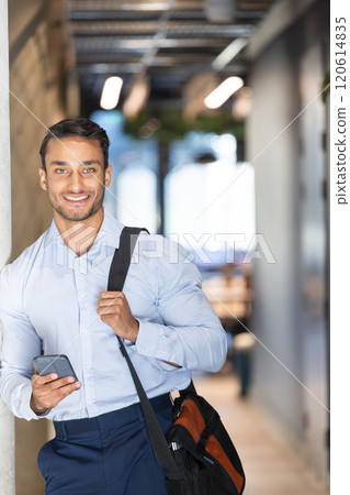 Smiling businessman holding smartphone and carrying bag in modern office hallway Smiling businessman holding smartphone and carrying bag in modern office hallway 120614835