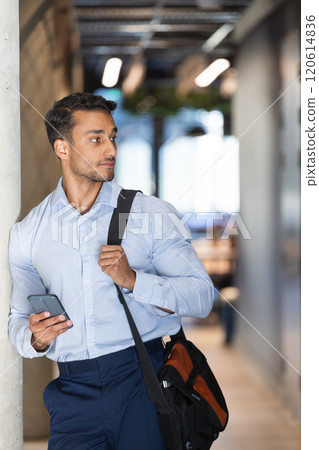 Holding smartphone, businessman with shoulder bag looking away in office hallway Holding smartphone, businessman with shoulder bag looking away in office hallway 120614836
