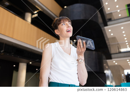 Speaking into smartphone, businesswoman smiling confidently in modern office building Speaking into smartphone, businesswoman smiling confidently in modern office building 120614863