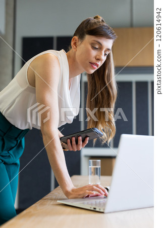 Using laptop and holding smartphone, businesswoman working in modern office Using laptop and holding smartphone, businesswoman working in modern office 120614942