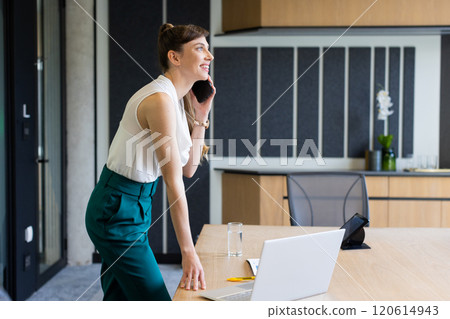 Talking on smartphone, woman standing at desk with laptop in modern office, copy space 120614943
