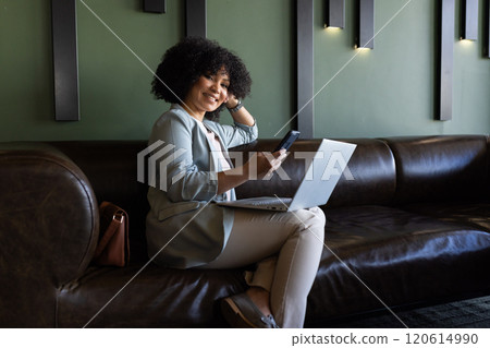 Using smartphone and laptop, businesswoman working remotely on leather couch 120614990