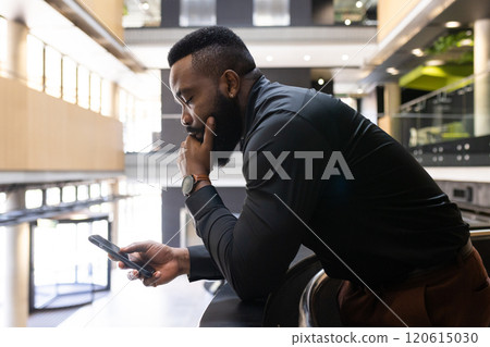 Leaning on railing, african american businessman using smartphone and thinking in modern office 120615030