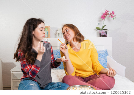 Two young women are sitting on the bed, eating popcorn and writing something down. The concept of quarantine, work from home, security and isolation Two young women are sitting on the bed, eating popcorn and writing something down. The concept of quarantine, work from home, security and isolation 120615362