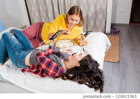 Two young women are lying on the bed, eating popcorn, using smarfton, and decorative cosmetics are lying next to them. Copy space. Concept of LGBT relationships and recreation at home Two young women are lying on the bed, eating popcorn, using smarfton, and decorative cosmetics are lying next to them. Copy space. Concept of LGBT relationships and recreation at home 120615363