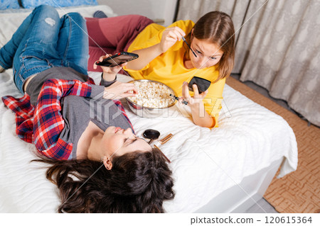 Two young women are lying on the bed, eating popcorn, using smarfton, and decorative cosmetics are lying next to them. Top view.Concept of LGBT relationships and recreation at home 120615364