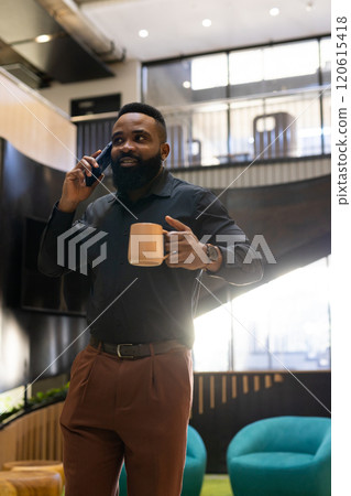 Talking on smartphone, African american man holding coffee cup in modern office setting 120615418