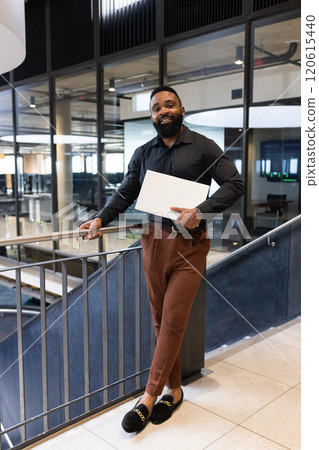 Holding laptop, African American businessman standing confidently in modern office building lobby Holding laptop, African American businessman standing confidently in modern office building lobby 120615440