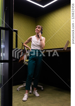 Talking on smartphone, woman standing in office with laptop on desk 120615456