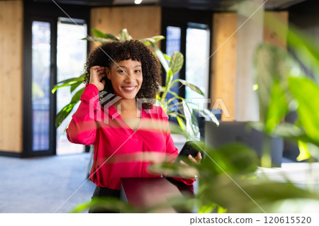 Holding smartphone and adjusting earpiece, smiling woman in office surrounded by plants Holding smartphone and adjusting earpiece, smiling woman in office surrounded by plants 120615520