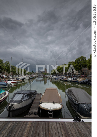 Serene harbor with moored boats under a dramatic cloudy sky. Quaint historic buildings and trees line the waterfront, reflecting in the calm green water, creating peaceful scene. Dordrecht landscape. Serene harbor with moored boats under a dramatic cloudy sky. Quaint historic buildings and trees line the waterfront, reflecting in the calm green water, creating peaceful scene. Dordrecht landscape. 120615639