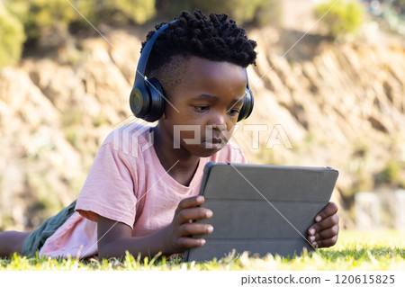 Using tablet and wearing headphones, african american boy studying outdoors on grass 120615825