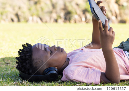 Lying on grass, african american boy using tablet and wearing headphones, enjoying outdoor learning 120615837